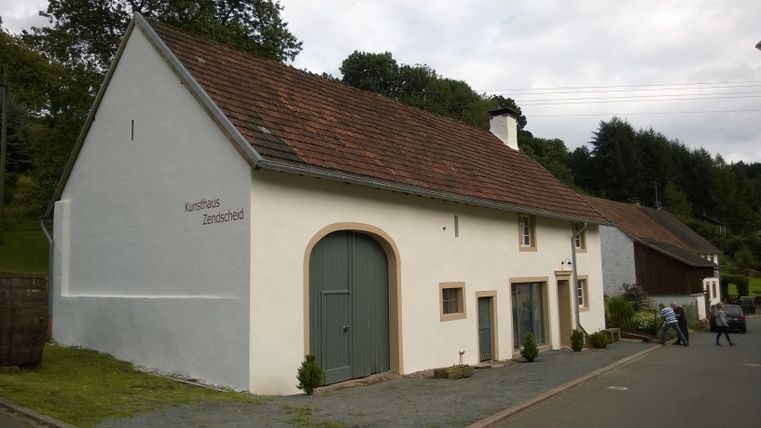 Ein weißes Gebäude mit der Aufschrift 'Kunsthaus Zendscheid' und einem grünen Tor, umgeben von Bäumen und einer Straße.