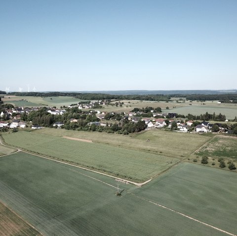 Vue aérienne d'Idenheim : une agglomération rurale entourée de champs verdoyants, avec des éoliennes à l'horizon sous un ciel dégagé., © Ingrid Penning