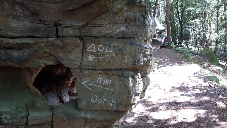 Eine Felsformation im Wald mit unleserlichen Zeichen und Pfeilen. Der Weg führt durch eine grüne Umgebung.