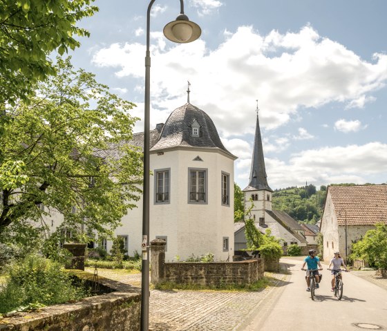Zwei Radfahrer fahren auf einer sonnigen Straße in Wolsfeld. Links stehen historische Gebäude und ein Baum, im Hintergrund ist eine Kirche zu sehen., © Eifel Tourismus GmbH, Dominik Ketz