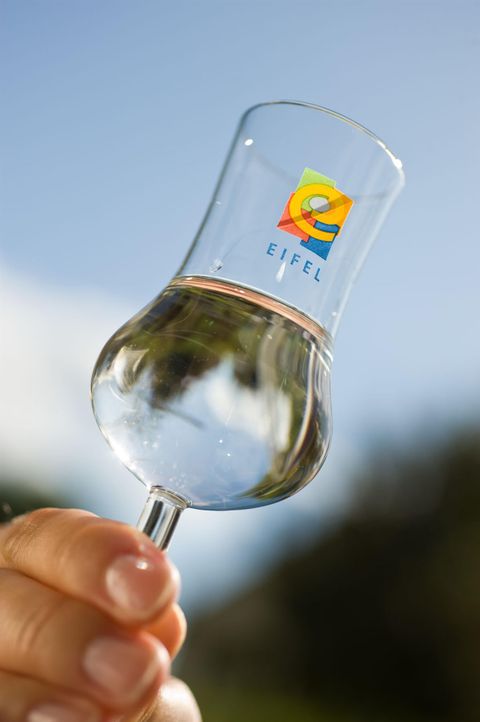 Ein transparentes Glas mit Wasser wird von einer Hand gehalten. Das Glas trägt ein buntes Logo mit dem Namen "Eifel".