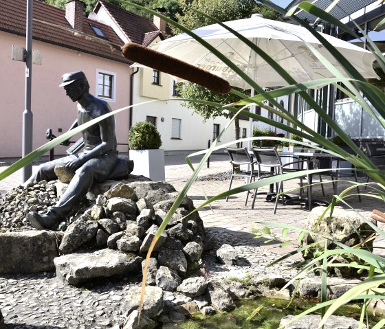 Bronzen beeld van een man op stenen, omgeven door riet en een kleine vijver. Huizen en een parasol zijn te zien op de achtergrond., © Tourist-Information Bitburger Land