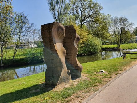 Steinskulptur am Flussufer mit Bäumen im Hintergrund.