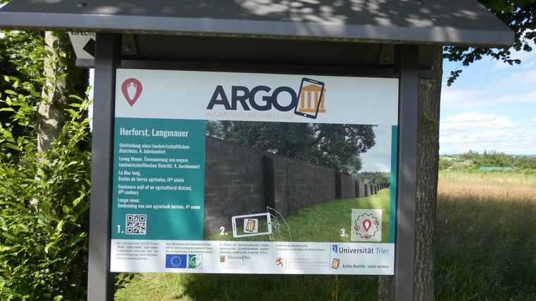 Ein Schild mit Informationen über das ARGO III Projekt in Herford. Im Hintergrund ist eine historische Mauer zu sehen, umgeben von Wiese und Bäumen.