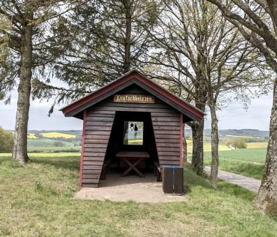 Een kleine houten hut met het bordje 'Knutschheisjen' staat tussen bomen in een groen landschap van velden en heuvels., © A. Girards