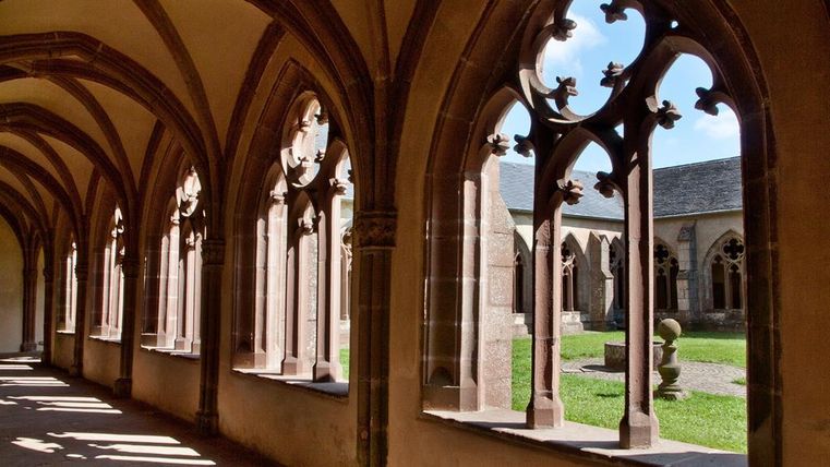 Ein Kreuzgang mit gotischen Fenstern und einem grünen Innenhof. Das Licht fällt sanft durch die Bögen und schafft eine ruhige Atmosphäre.