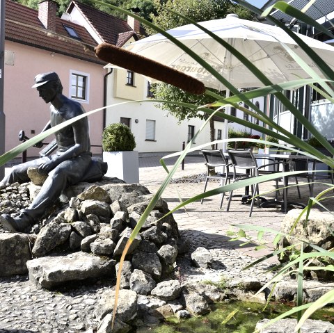 Bronzestatue eines Mannes auf Steinen, umgeben von Schilf und einem kleinen Teich. Im Hintergrund sind Häuser und ein Sonnenschirm zu sehen., © Tourist-Information Bitburger Land