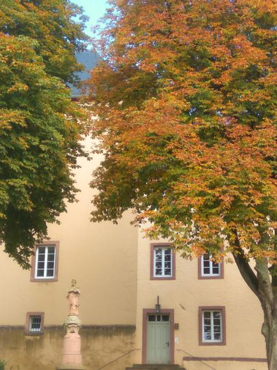 Ein historisches Gebäude mit gelber Fassade und bunten Bäumen im Herbst. Im Vordergrund steht eine kleine Statue.