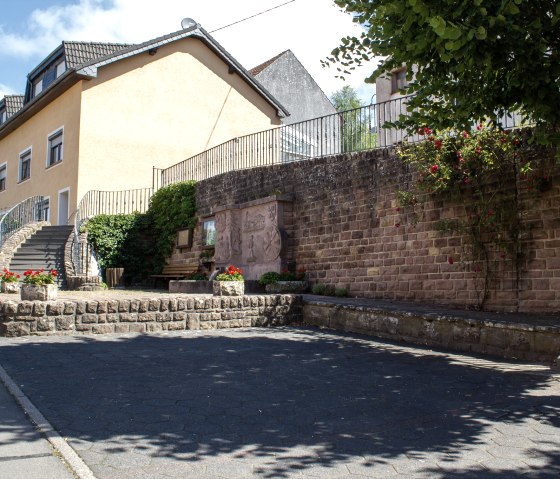 Gepflasterter Parkplatz vor einem gelben Geb&auml;ude mit Treppe. Blumenbeete und eine Steinmauer mit Relief. Baum spendet Schatten., &copy; TI Bitburger Land M. Mayer