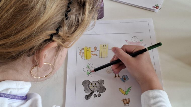A child is drawing with a green marker on a sheet of paper. The drawing shows an elephant and various colorful motifs.