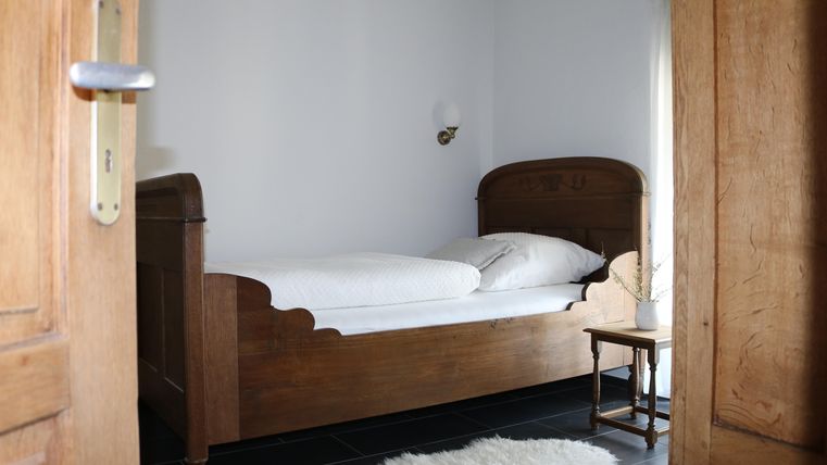Een eenvoudige slaapkamer met een houten bed en witte beddengoed. Naast het bed staat een kleine tafel en een tapijt ligt op de vloer.