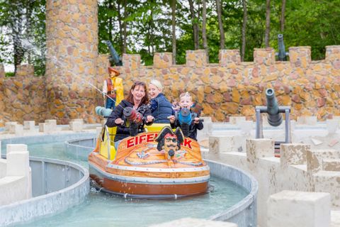 Eine fröhliche Familie fährt mit einem Wasserboot in einem Vergnügungspark. Im Hintergrund sind eine Burgmauer und Bäume zu sehen.