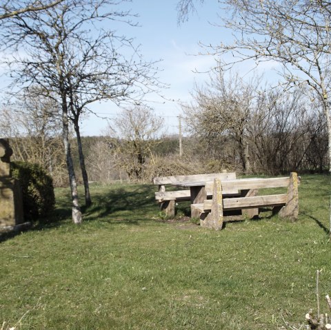 Aire de repos avec bancs en bois et croix en pierre sur une prairie, entourée d'arbres dénudés sous un ciel bleu., © M. Bach