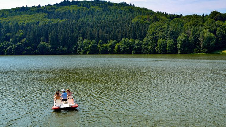 Ein Ruderboot mit drei Personen auf einem ruhigen See. Im Hintergrund sind bewaldete Hügel zu sehen.