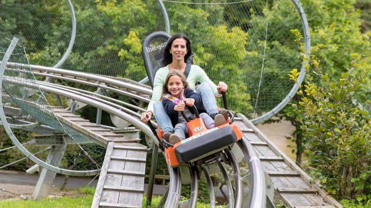 Eine Mutter fährt mit ihrem Kind auf einer Rodelbahn. Sie lachen und genießen die Fahrt inmitten von grünen Bäumen.