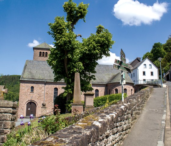 Die Kirche St. Maximin in Kyllburg, umgeben von blühenden Blumen und einem Wegweiser, bei sonnigem Wetter., © TI Bitburger Land