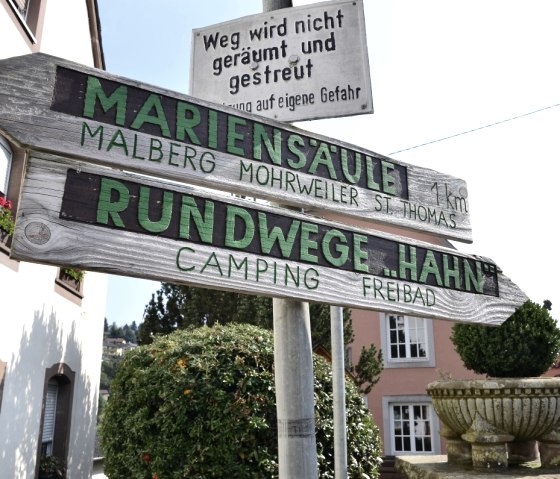 Ein hölzerner Wegweiser in Kyllburg zeigt Richtungen zur Marien-Säule und Rundwege Hahn. Im Hintergrund sind Gebäude und Pflanzen zu sehen., © TI Bitburger Land
