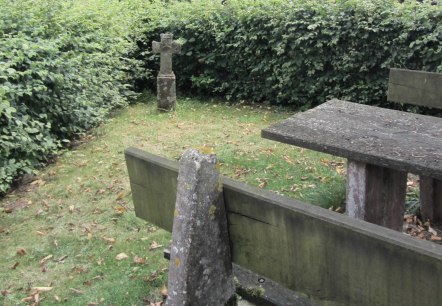 Ein Steinkreuz steht auf einer Wiese neben einem verwitterten Holztisch, umgeben von dichten Hecken und Bäumen., © M. Bach