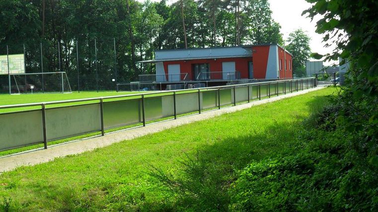Ein Fußballplatz mit grünem Rasen und einem modernen roten Gebäude im Hintergrund. Umgeben von Bäumen und einem gepflegten Weg.