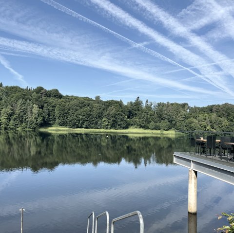 Der Stausee Bitburg mit ruhigem Wasser, umgeben von Wald. Eine Terrasse mit Tischen und Sonnenschirm ragt ins Bild. Klare Himmel mit Kondensstreifen., &copy; TI Bitburger Land