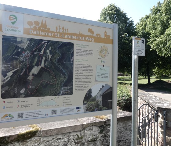 Informationstafel des Dahlemer St.-Lambertus-Wegs mit Karte und Infos, umgeben von Bäumen und einem Weg im Hintergrund., © TI Bitburger Land