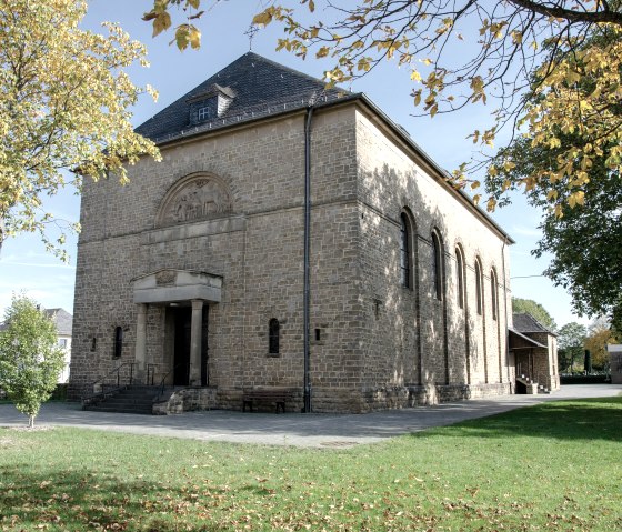 De Sint-Hubertuskerk in Wolsfeld, gebouwd van lichtgekleurde steen, staat in een herfstachtige omgeving met bomen en groene gazons., © Tourist-Information Bitburger Land