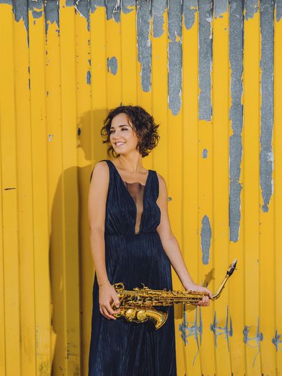 A smiling woman in a black dress is holding a saxophone. The background is a bright yellow wall.