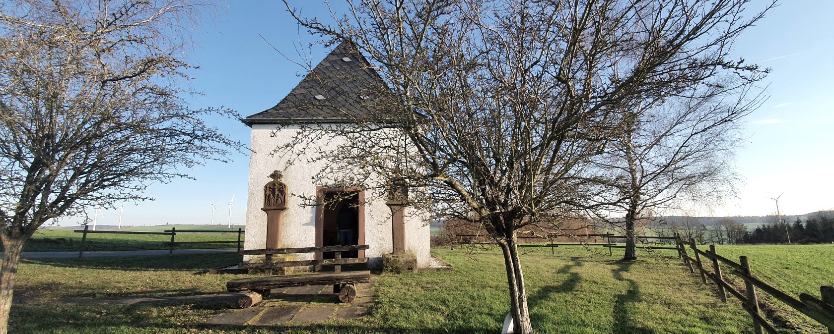 Une petite chapelle sur une prairie verte, entourée d'arbres dénudés. En arrière-plan, on aperçoit des éoliennes., © TI Bitburger Land
