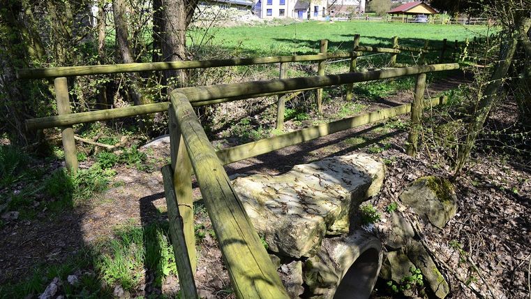 Holzgeländer und Steine über einem kleinen Bach, im Hintergrund ein blaues Haus und grüne Wiese.