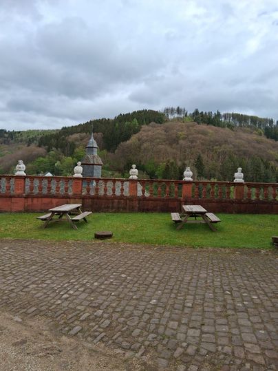 Eine Aussicht auf eine grüne Landschaft mit sanften Hügeln im Hintergrund. Im Vordergrund befinden sich einige Holztische auf einem gepflasterten Boden.