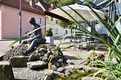Bronzestatue eines sitzenden Mannes auf Steinen, umgeben von Schilf und einem kleinen Teich, mit Häusern und einem Sonnenschirm im Hintergrund.