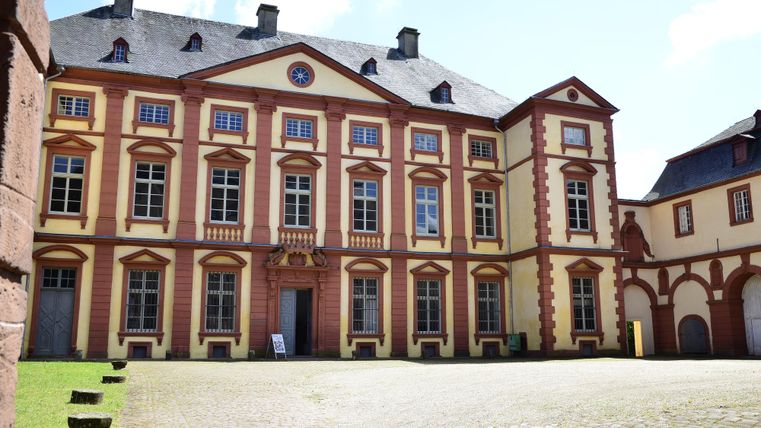 Un bâtiment historique avec des façades colorées et de grandes fenêtres. La cour est pavée et bien éclairée par le soleil.