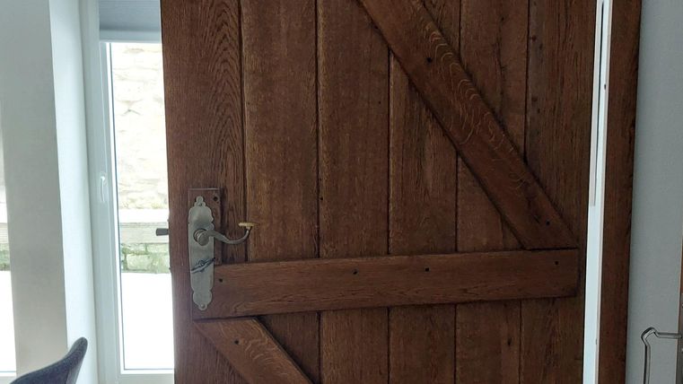 Een brown houten deur met een rustieke uitstraling staat open. Op de achtergrond zijn meubels en een raam zichtbaar.