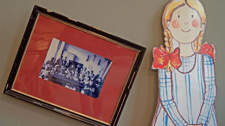 Ein gerahmtes Foto einer Gruppe von Kindern in einem Klassenzimmer hängt an der Wand. Neben dem Bild steht eine gemalte Figur eines Mädchens mit Zöpfen und einer Schleife.