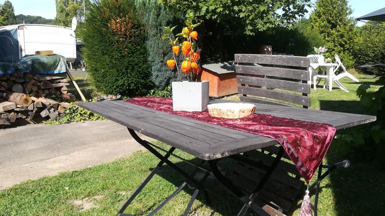 Une table en bois simple avec une nappe rouge est installée à l'extérieur. Sur la table se trouve un vase de fleurs et en arrière-plan, on peut voir des arbres et d'autres meubles de jardin.