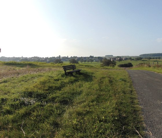 Eine Holzbank steht auf einer grünen Wiese neben einem Weg. Im Hintergrund ist ein Dorf zu sehen, umgeben von Feldern und Bäumen., © Conny Meier