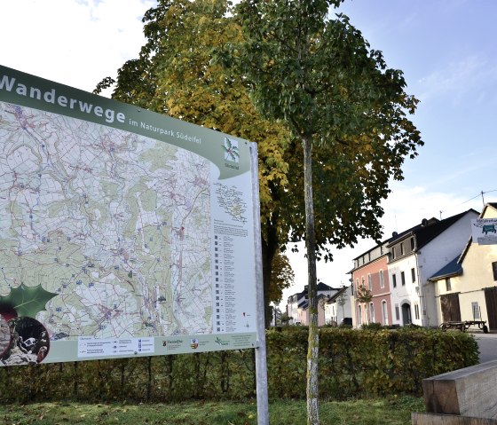 Wanderkarte im Naturpark Südeifel vor herbstlichen Bäumen und Häusern in Wolsfeld. Eine Ruhebank und ein Briefkasten sind ebenfalls zu sehen., © TI Bitburger Land