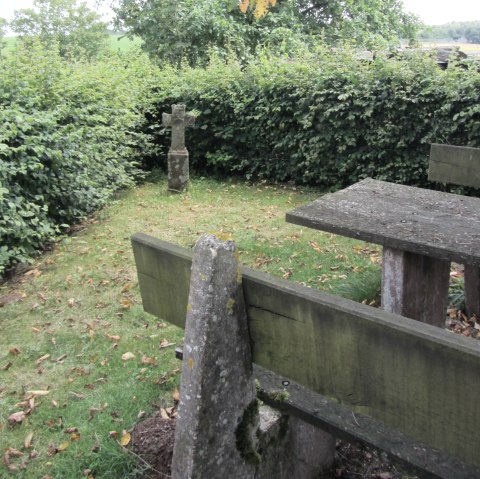 Een stenen kruis staat op een weiland naast een verweerde houten tafel, omringd door dichte heggen en bomen., © M. Bach