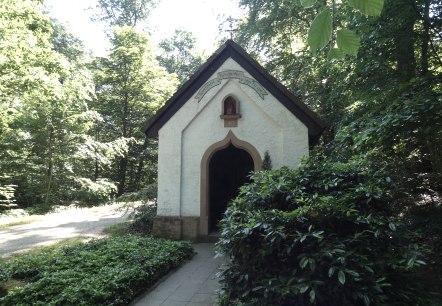 Eine kleine Kapelle aus dem Jahr 1877 steht im Wald, umgeben von Bäumen und einem Weg. Die Sonne scheint durch das Blätterdach.