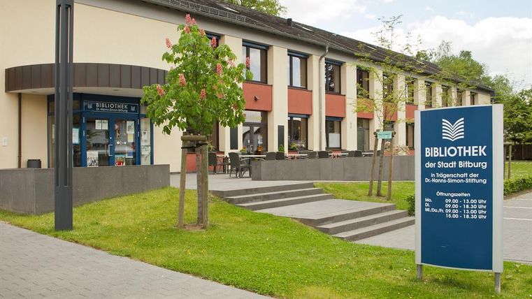 Ein modernes Bibliotheksgebäude mit einem einladenden Eingang. Auf dem Gelände sind Bäume und eine Infotafel mit Öffnungszeiten zu sehen.