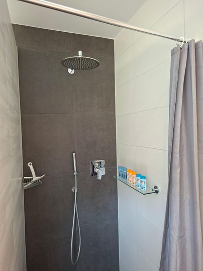Une douche moderne avec des carreaux gris et une grande douche pluie. Sur une étagère en verre se trouvent différents produits de soin.