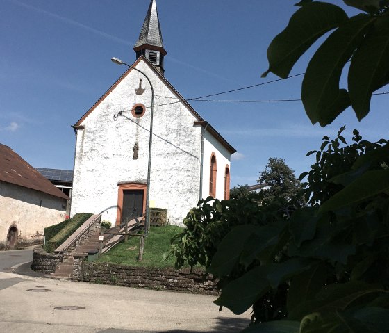 Kleine Kapelle mit spitzem Turm und orange umrahmten Fenstern in Etteldorf, umgeben von Bäumen und blauem Himmel., © Tourist-Info Bitburger Land M.Mayer