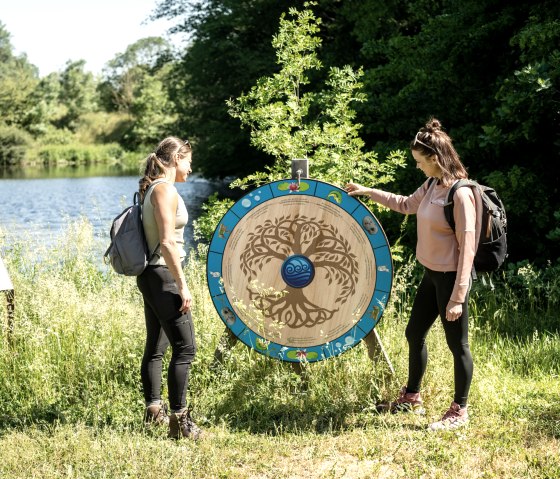 Deux femmes portant des sacs à dos contemplent une roue en bois décorative au bord d'un lac, entourée d'une nature verdoyante., © Eifel Tourismus GmbH, Dominik Ketz