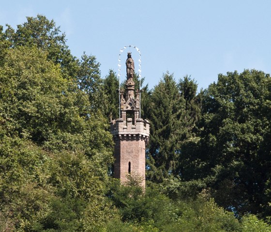 De Mariazuil in Kyllburg rijst op uit het dichte bos. Een standbeeld staat op een stenen toren, omringd door groene bomen en een blauwe lucht., © TI Bitburger Land