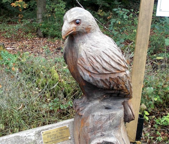 A wooden sculpture of a bird sits on a tree trunk in the forest. A plaque is attached to the sculpture., © Tourist-Information Bitburger Land