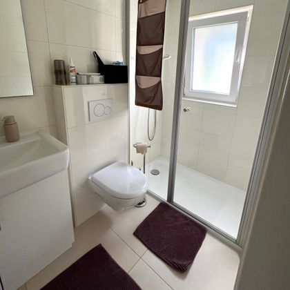 Ein modernes Badezimmer mit Dusche, Toilette und Waschbecken. Es sind zwei violette Badeteppiche auf dem Boden platziert.