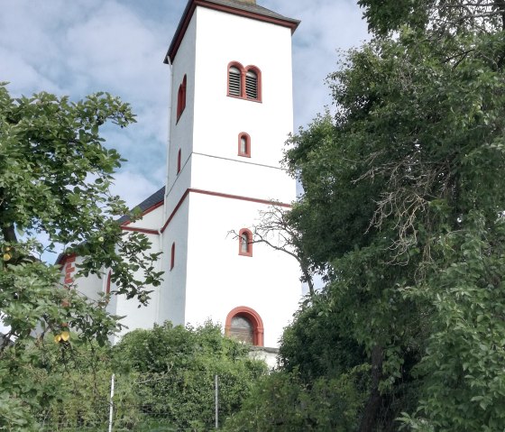 Witte kerk met rood dak en toren, omringd door groene bomen en blauwe lucht op de achtergrond., © TI Bitburger Land