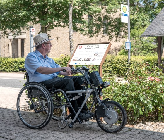 Un homme en fauteuil roulant lit un panneau du sentier thématique Baukultur Eifel à Wolsfeld. En arrière-plan, on aperçoit des arbres et un bâtiment., © Naturpark Südeifel, Thomas Urbany