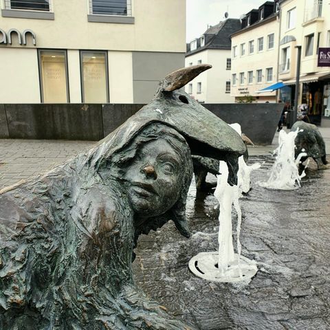 Eine kunstvolle Bronzeskulptur, die eine Figur mit einem Tierkopf darstellt. Im Hintergrund sind kleine Wasserfontänen zu sehen, die der Szene Leben verleihen.