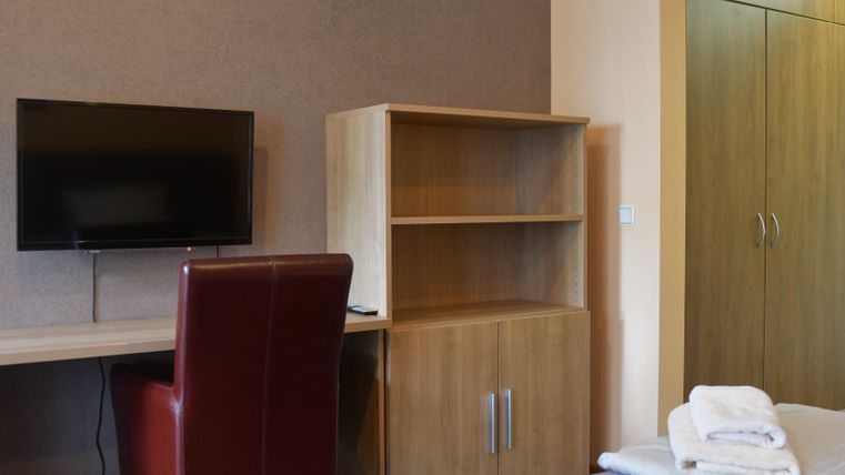 Ein modernes Schlafzimmer mit einem Schreibtisch, einem Stuhl und einem Fernseher. Im Hintergrund befindet sich ein Kleiderschrank und ein Bett mit frischen Handtüchern.